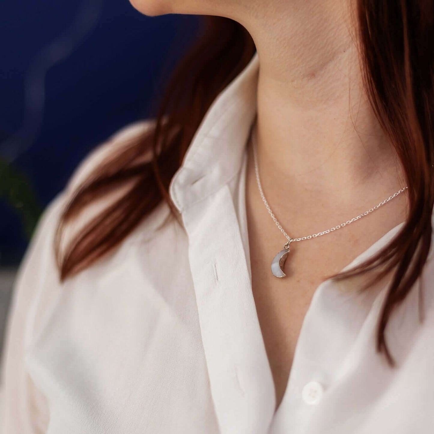 Close-up of a person wearing a white shirt and a Made Here with Love Moon Agate Charm Necklace, showcasing its delicate crescent moon pendant. The person's long hair is partially visible, while the out-of-focus background draws attention to the necklace and shirt. Perfectly packaged in an elegant gift box, this piece makes an ideal present for any occasion.