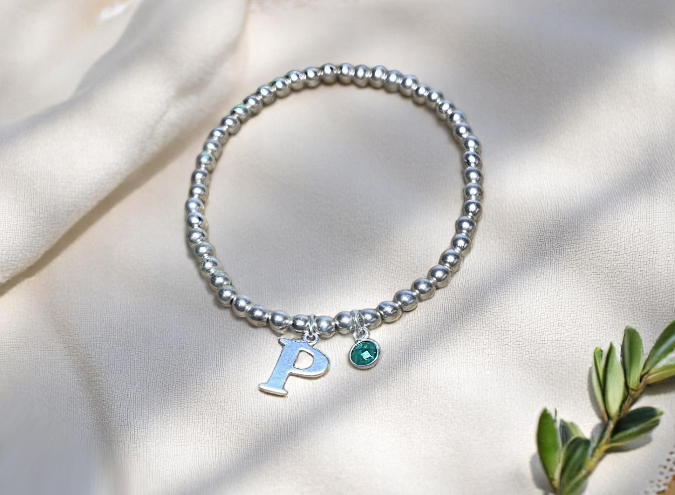 Sterling silver personalised bracelet with emerald May birthstone charm and initial letter pendant – resting on fabric with botanical elements.