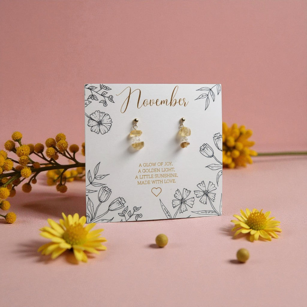 November citrine birthstone earrings displayed on a floral illustrated card reading ‘A glow of joy, a golden light, a little sunshine. Made with love.’ The earrings and card are surrounded by yellow flowers on a soft pink background.