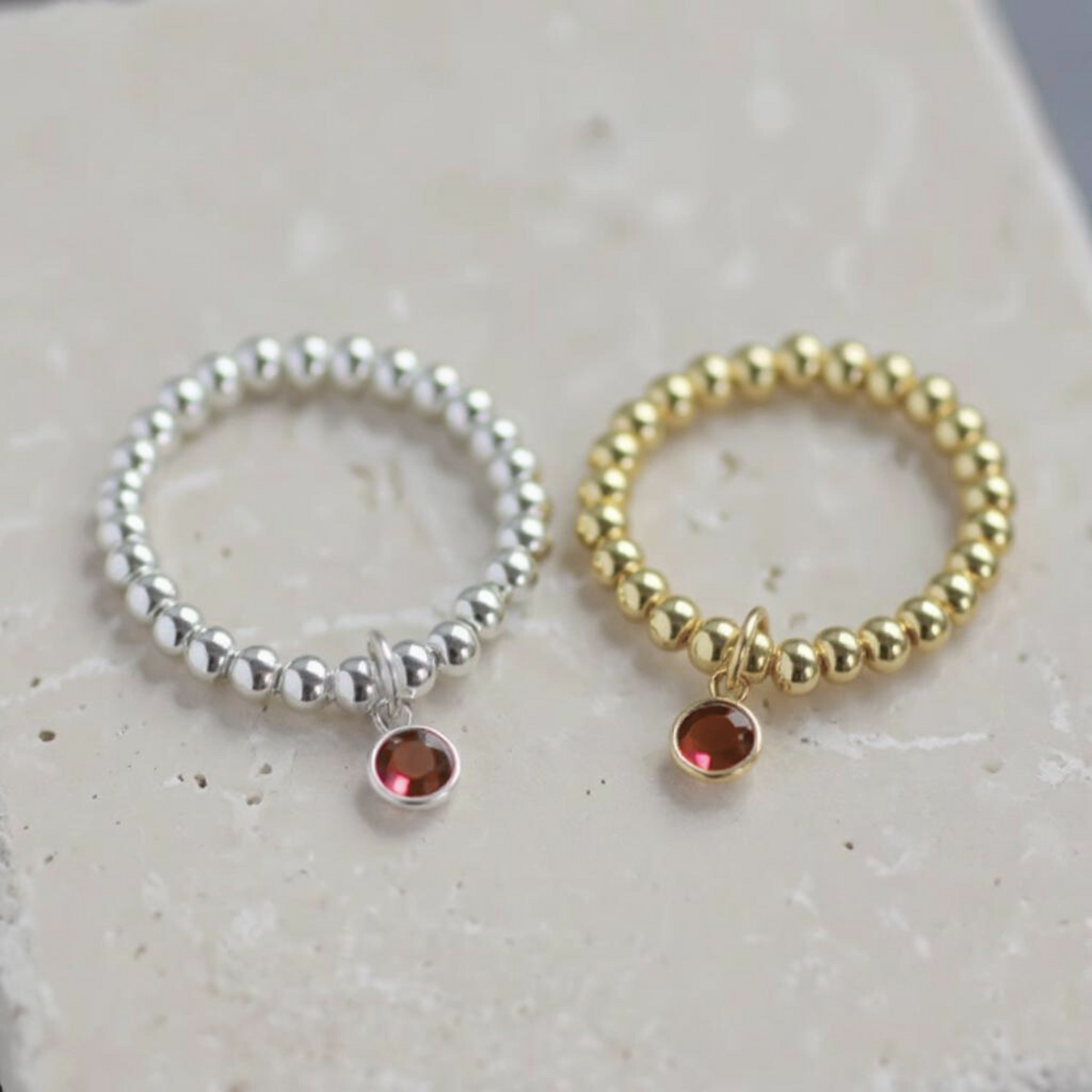 July birthstone stretch rings in silver and gold with ruby-coloured charms, displayed on neutral stone background, Made Here With Love.