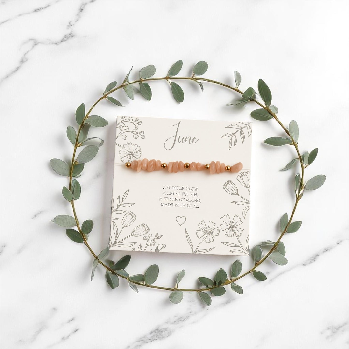 Peach moonstone June birthstone stretch bracelet with gold accents, displayed on a floral illustrated message card and surrounded by a eucalyptus wreath. A handmade crystal bracelet and meaningful gift for her, styled on a marble background.