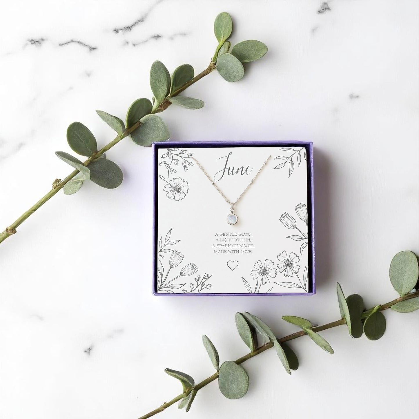 Handmade June birthstone necklace with a delicate moonstone charm on a fine chain, displayed in a purple gift box with floral message card. A meaningful crystal jewellery gift for her, styled with eucalyptus on a marble background.