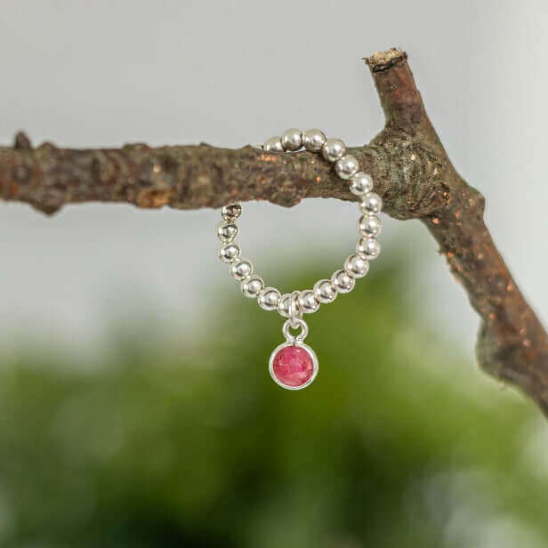 Sterling silver stretch bead ring with ruby birthstone charm, displayed hanging from a tree branch.