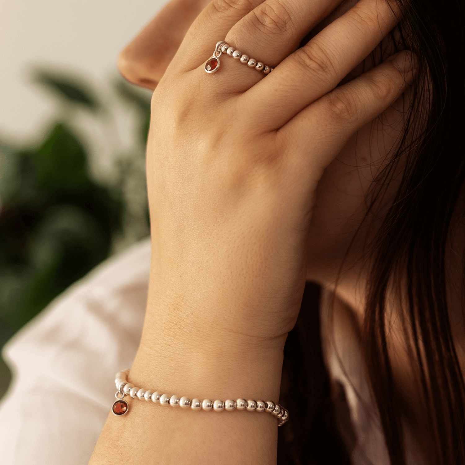  A person with long dark hair showcases a beaded bracelet and matching ring, each adorned with a small red garnet birthstone. Their hand is gently placed near their face, and the background is blurred with green foliage