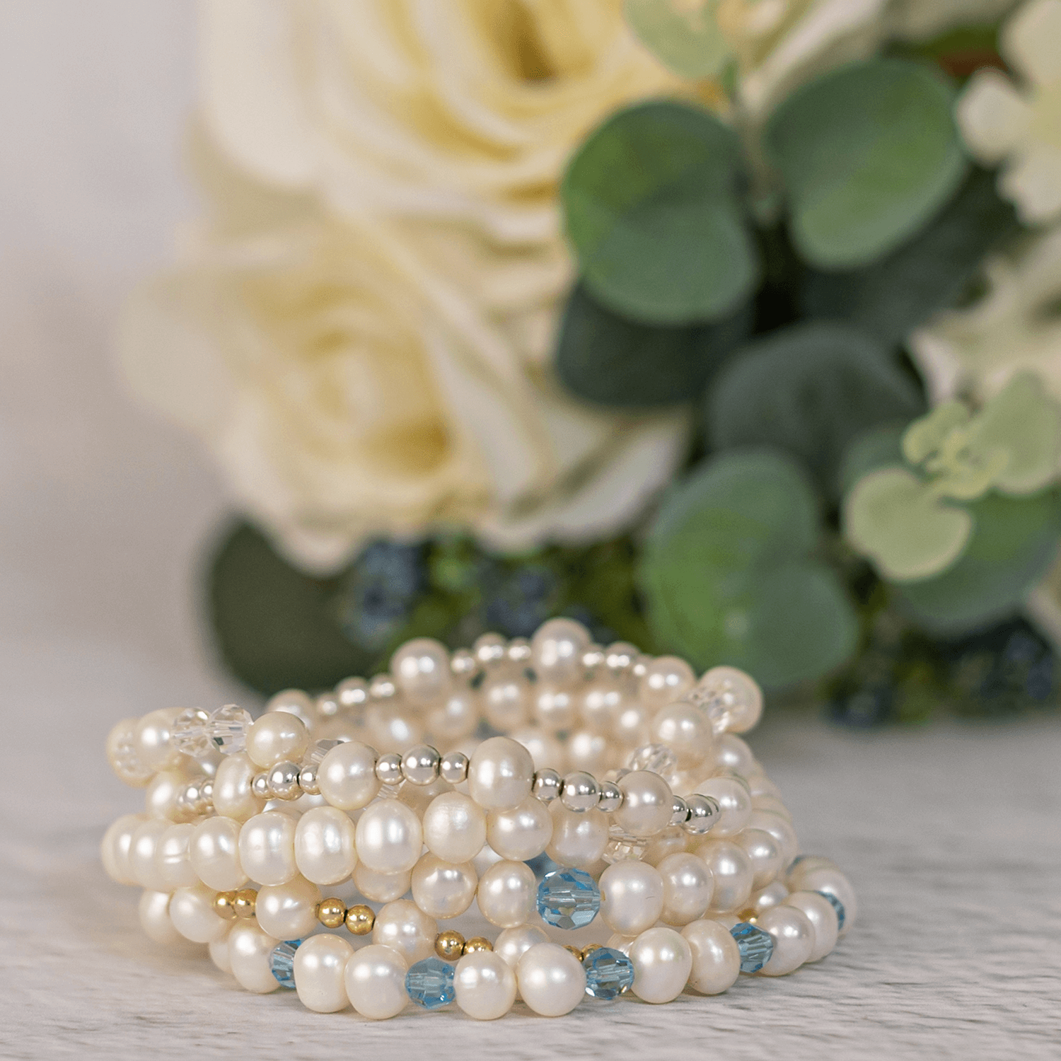  A beaded pearl bracelet perfect as bridal jewellery for a wedding, features alternating small white pearls, gold beads, and blue crystal beads elegantly sat. The bracelet rests on a light-colored surface with a backdrop of  green eucalyptus leaves.
