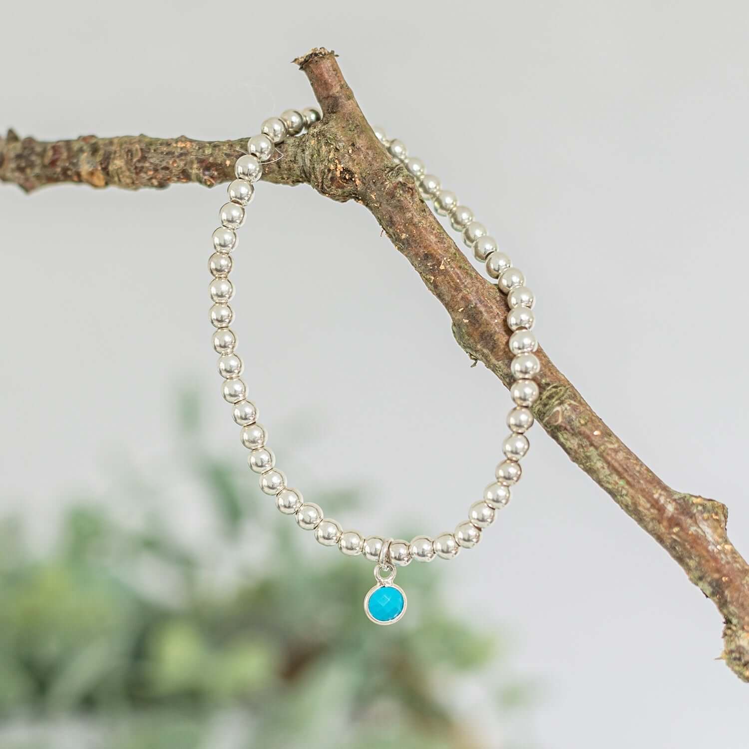  A beaded silver bracelet with a small turquoise charm, the December birthstone, hangs on a brown branch. The background is softly blurred with hints of green, giving a natural and serene vibe to the image
