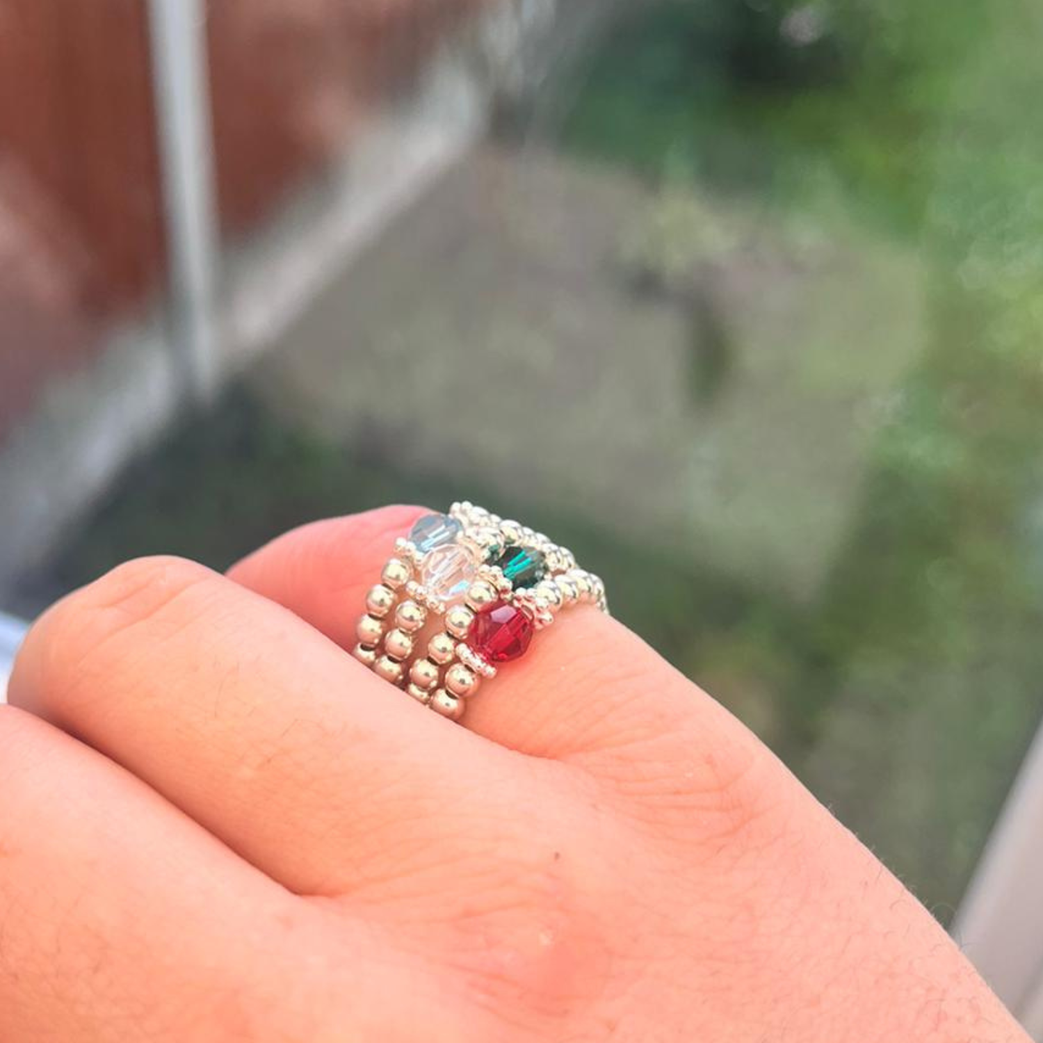 A close-up of a person's hand wearing a beaded ring adorned with various colored beads, including clear, green, and red. The background is slightly blurred, showing a window with a hint of greenery outside.