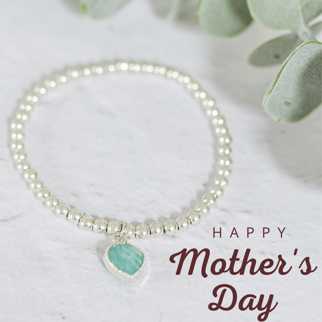 A silver beaded bracelet with a heart-shaped pendant is displayed. Green leaves are in the background. The text "Happy Mother's Day" is written in elegant maroon lettering in the bottom right corner of the image, perfect jewellery for Mother's Day