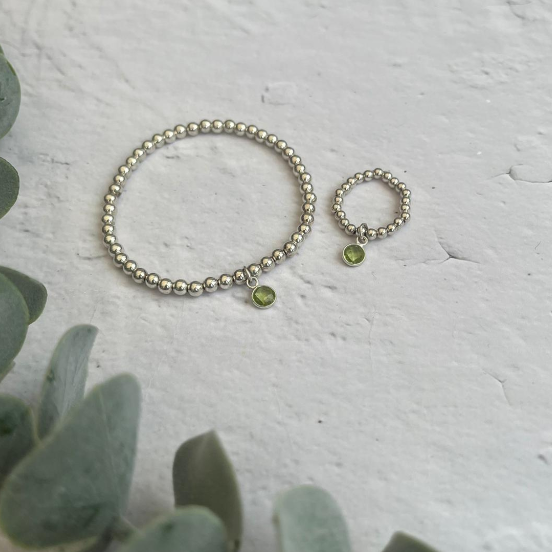 A close-up of a beaded silver ring and bracelet, each featuring a small green peridot charm. These pieces are part of matching jewellery sets. The rings are placed on a textured white surface, with green leafy plants in the left corner of the image.