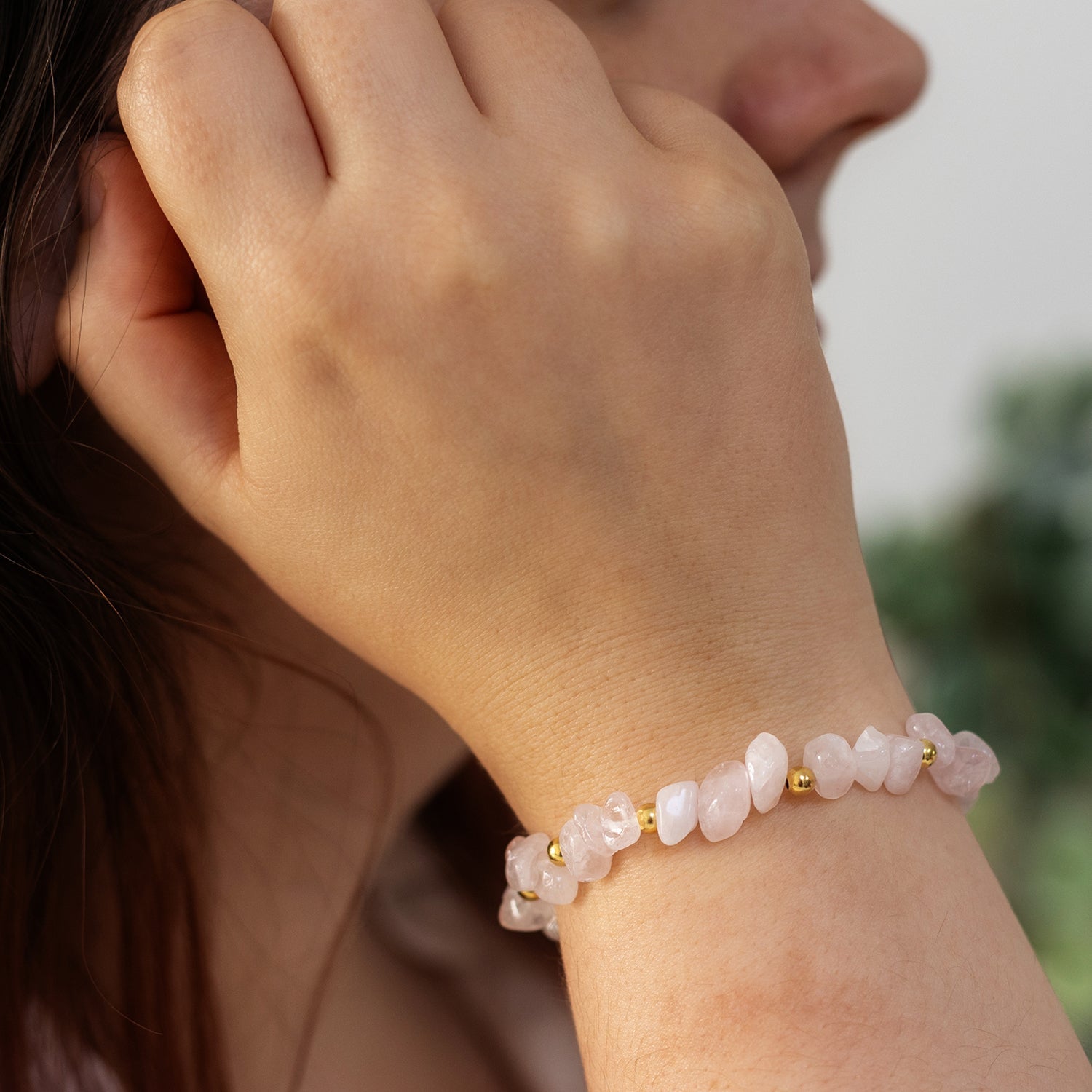 A person with long brown hair wears a bracelet made of irregularly shaped rose quartz beads. As they raise their hand near their face, the softly blurred greenery in the background highlights this exquisite piece of October birthstone jewellery