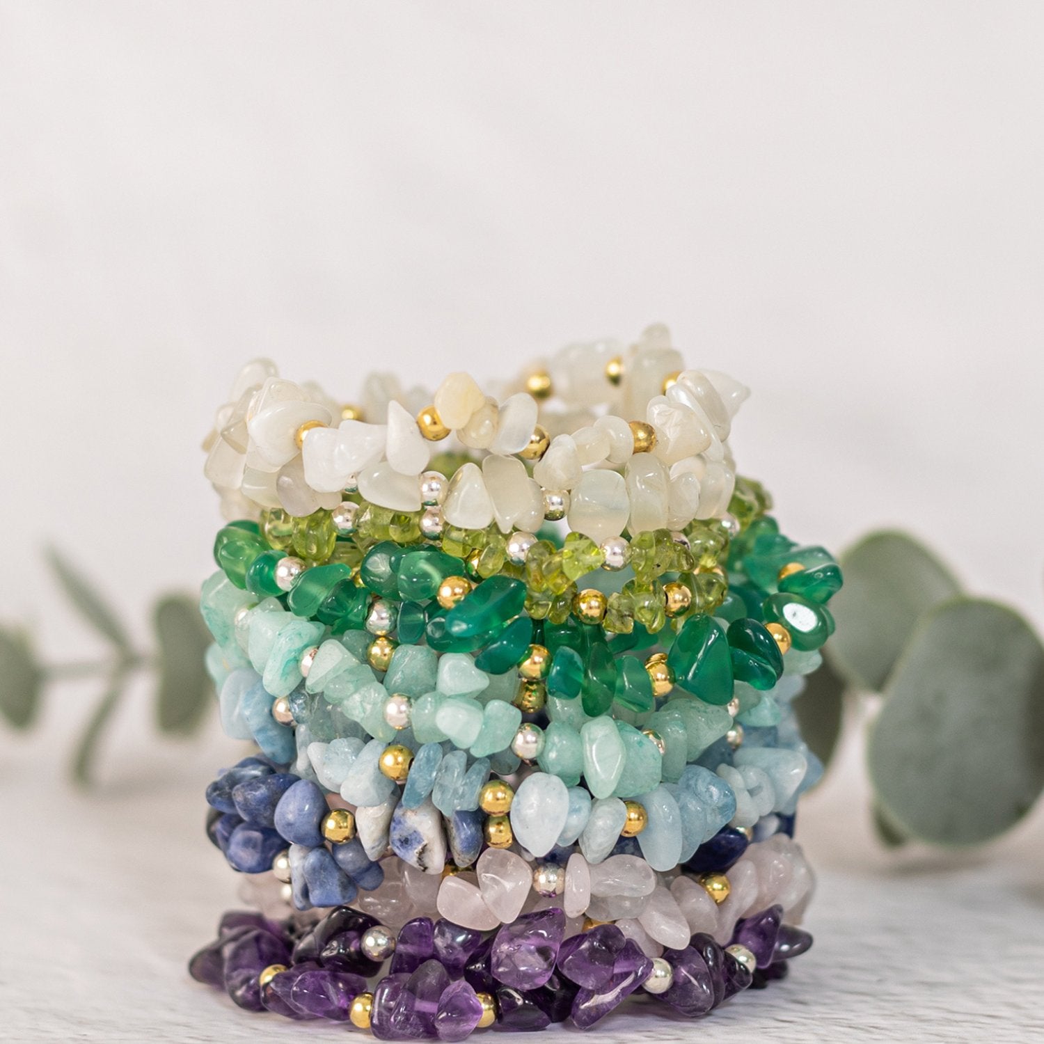  A stack of colorful gemstone bracelets made from various types of gemstones, including green, blue, white, and purple stones. The bracelets also feature small gold-colored beads and are displayed on a white surface with some greenery in the background.