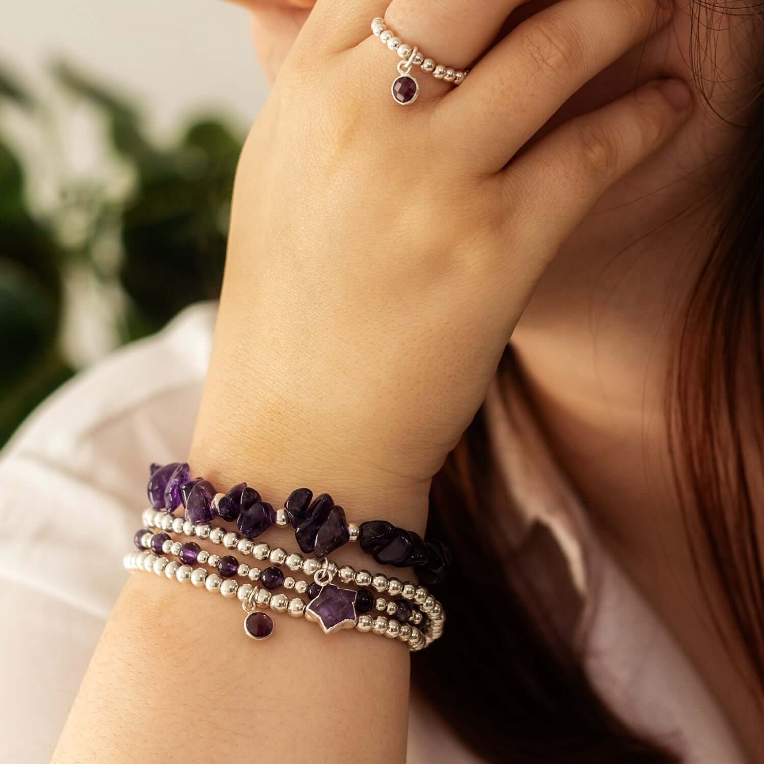 A close-up of a person wearing a silver ring with February birthstone jewellery, a purple gemstone, and a matching set of beaded bracelets with purple and silver beads. The person’s hand is near their face, against a backdrop of lush greenery