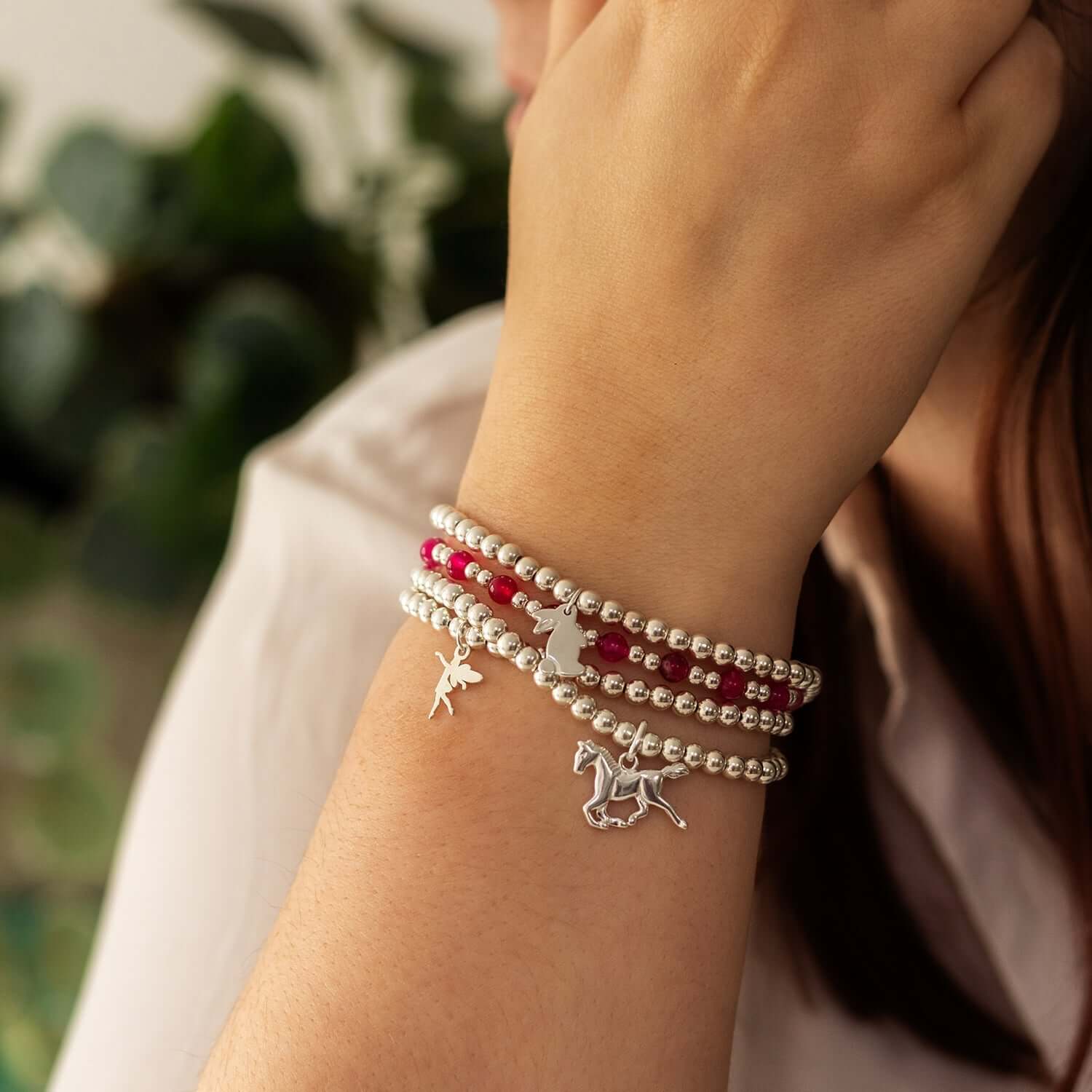  A person wearing a sterling silver charm bracelet adorned with small charms shaped like a fairy, a bunny, and a running horse. The bracelet features alternating red and silver beads. The person's arm is shown close-up with a blurred green background
