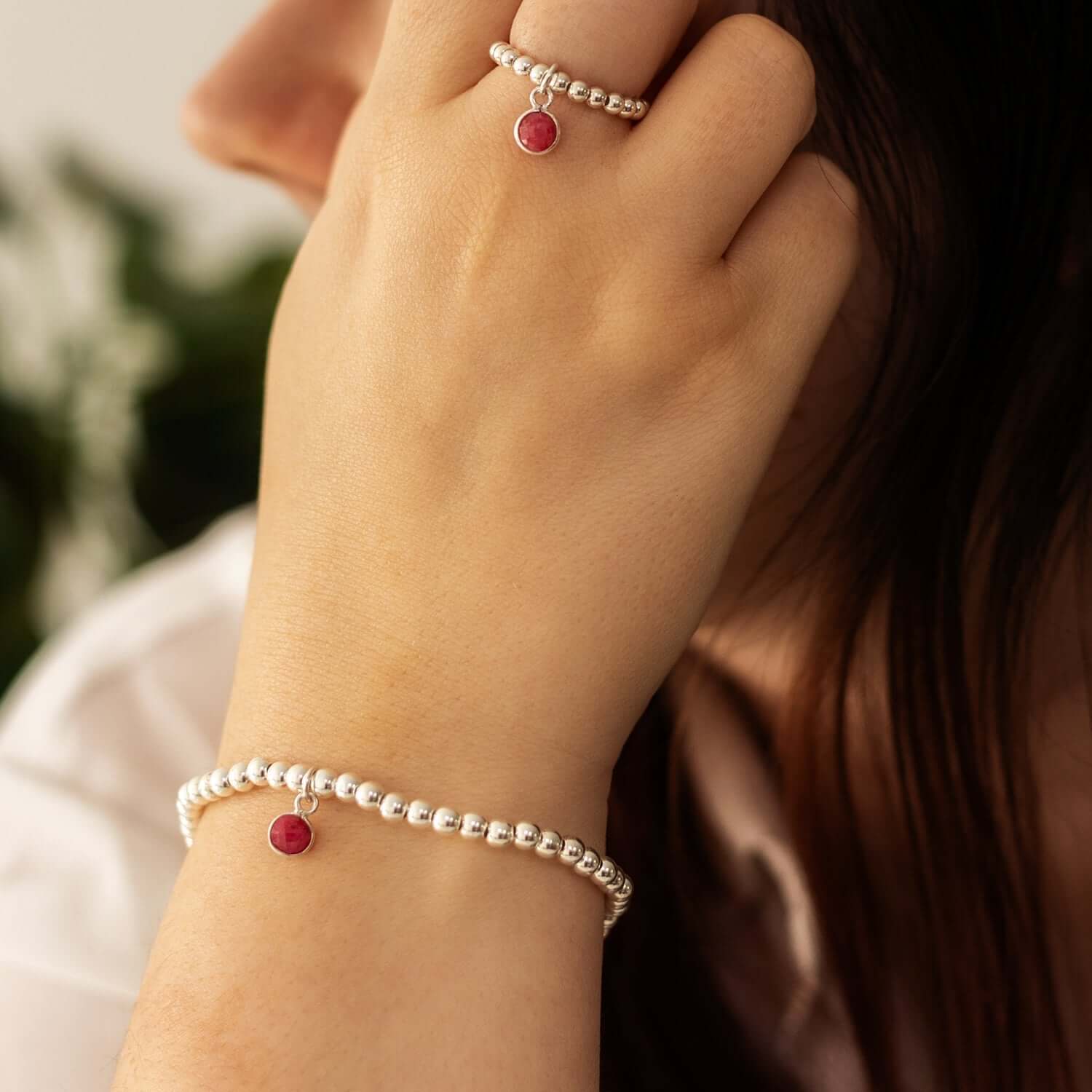   A close-up image of a person with their hand near their face, showcasing a matching beaded bracelet and ring. Both the bracelet and ring feature round, red gemstone charms, highlighting the elegance of ruby birthstone jewellery. 