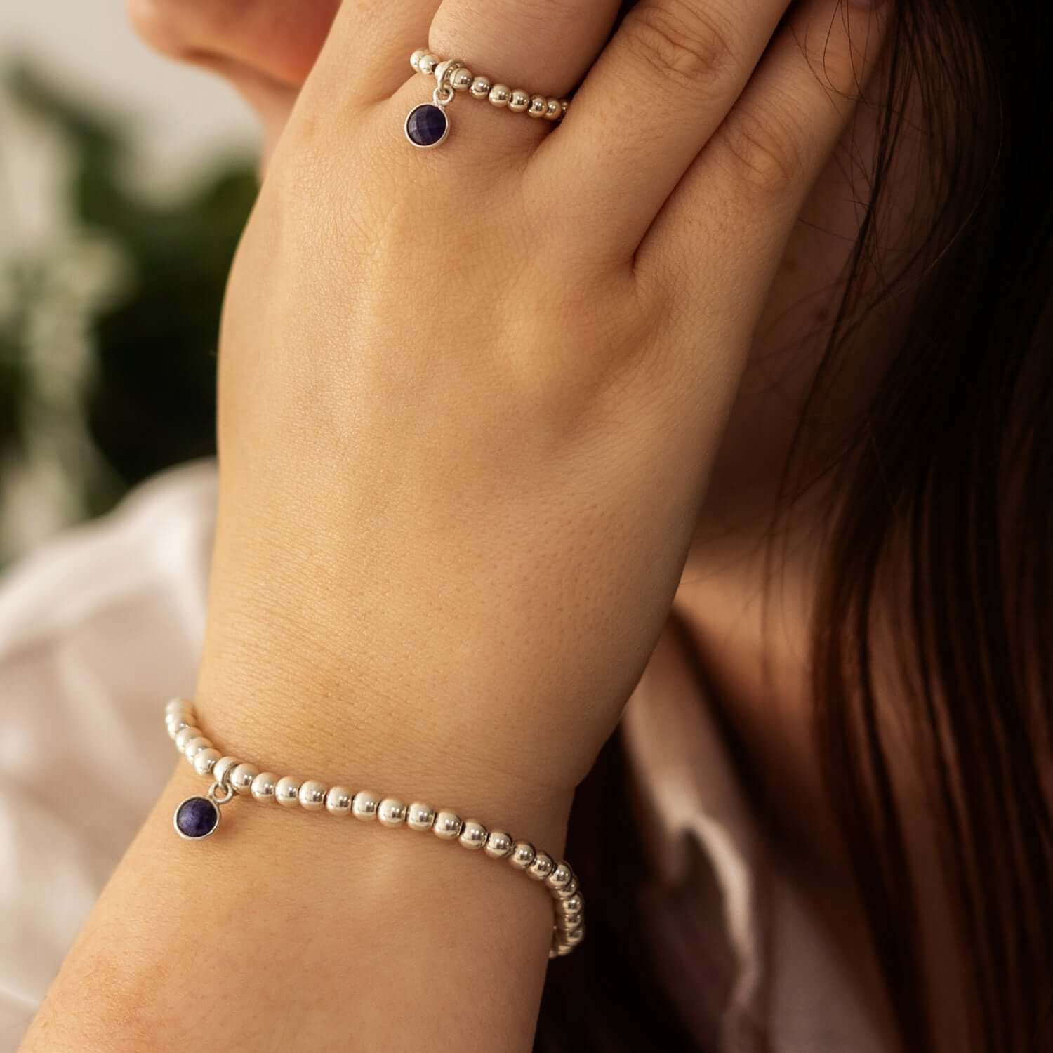 A person wearing a pearl bracelet and matching ring with small purple gemstone charms, reminiscent of September birthstone jewellery, holds their hand to their face. The background is blurred, with hints of greenery