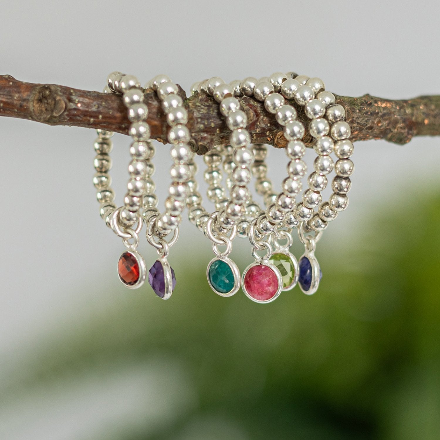 Six silver beaded rings, each with a colored gemstone charm in red, orange, purple, green, blue, and pink, are displayed on a brown branch against a blurred green background. These stackable birthstone rings add a personalized touch to your collection