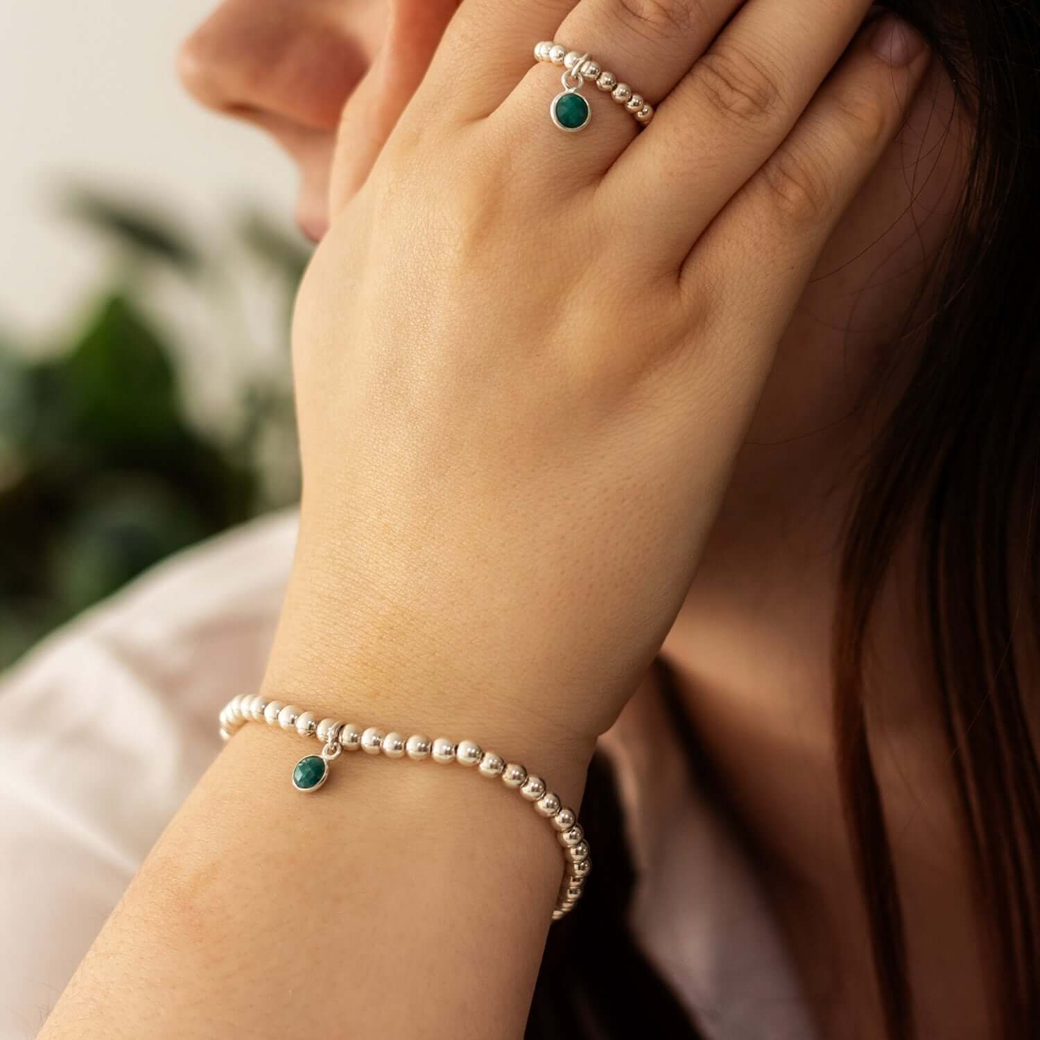 A close-up of a woman's hand resting on her cheek, showcasing May birthstone jewelry. She is wearing a silver beaded bracelet with a small green gemstone charm and a matching ring with a green stone. The background is softly blurred with hints of greenery