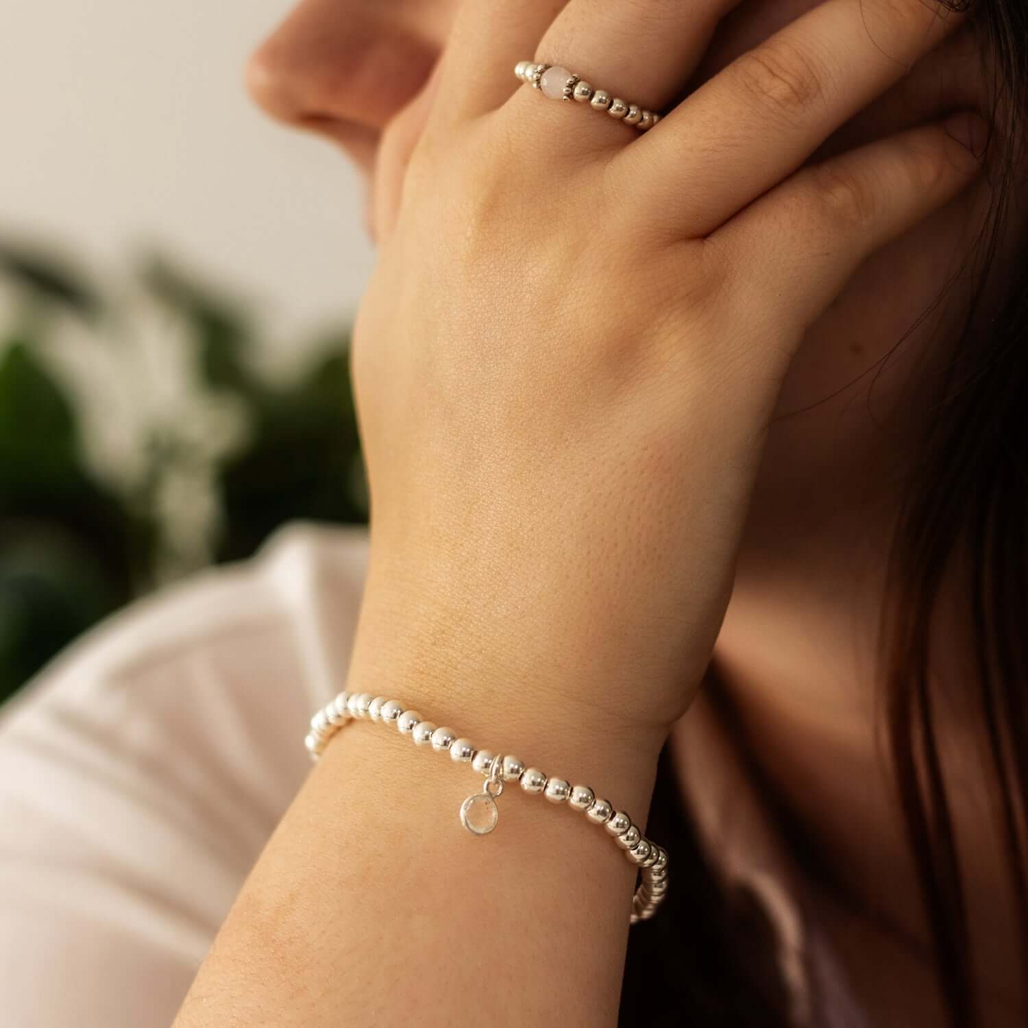A close-up of a person resting their hand on their face. They are wearing a beaded silver ring and a matching beaded silver bracelet with a small charm, reminiscent of April birthstone jewellery from the UK. The background is blurred with green foliage
