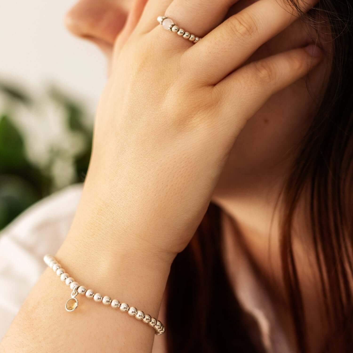 A close-up photo of a person wearing a bracelet on their wrist and a ring on their finger. Their hand touches the side of their face,with green plants blurred in the background, adding a serene touch to this November birthstone jewellery piece from the UK