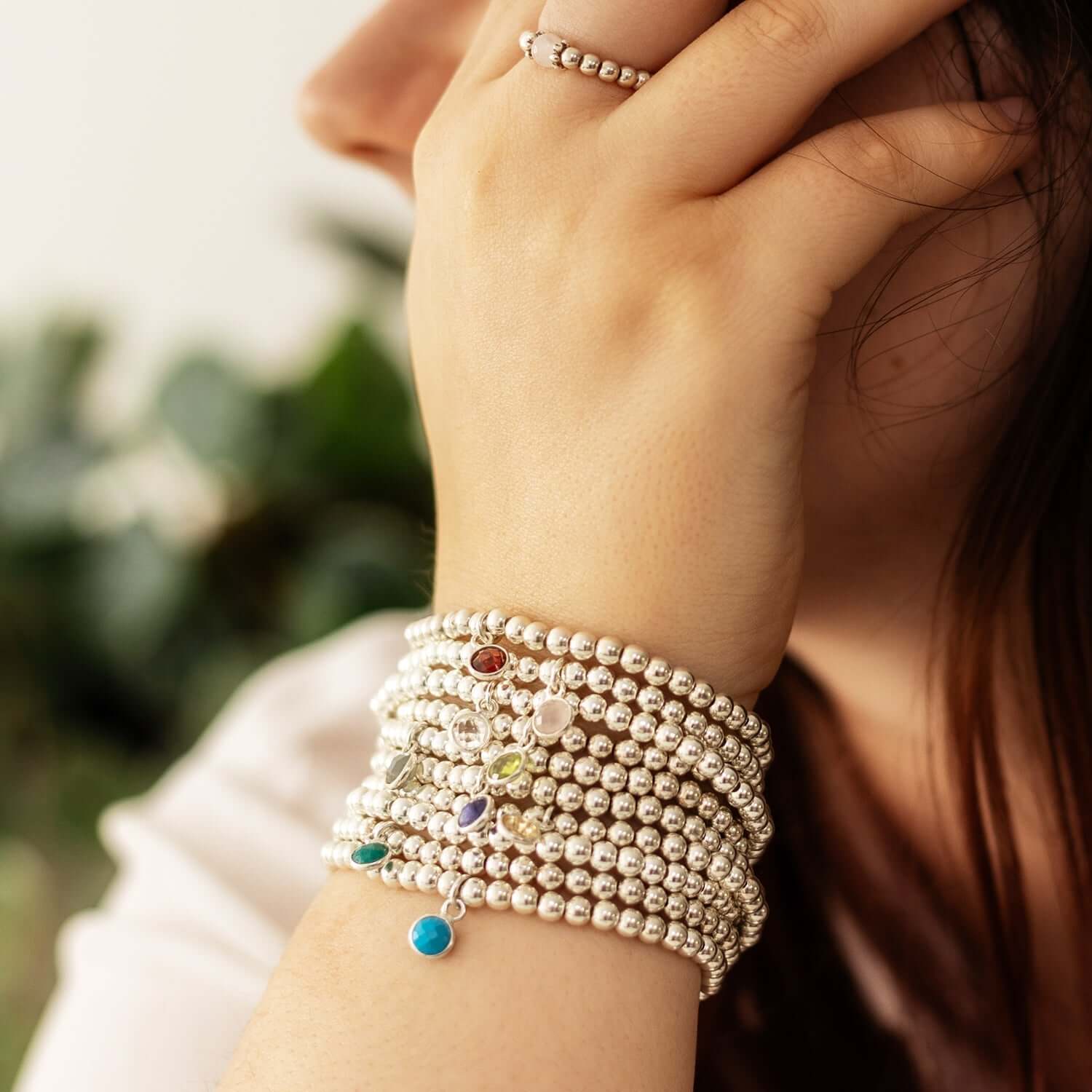 Close-up of a person with long hair, wearing multiple silver birthstone bracelets adorned with colorful charms on their wrist. The person has their hand lifted to their face, showing a silver beaded ring. A blurred green plant is in the background.