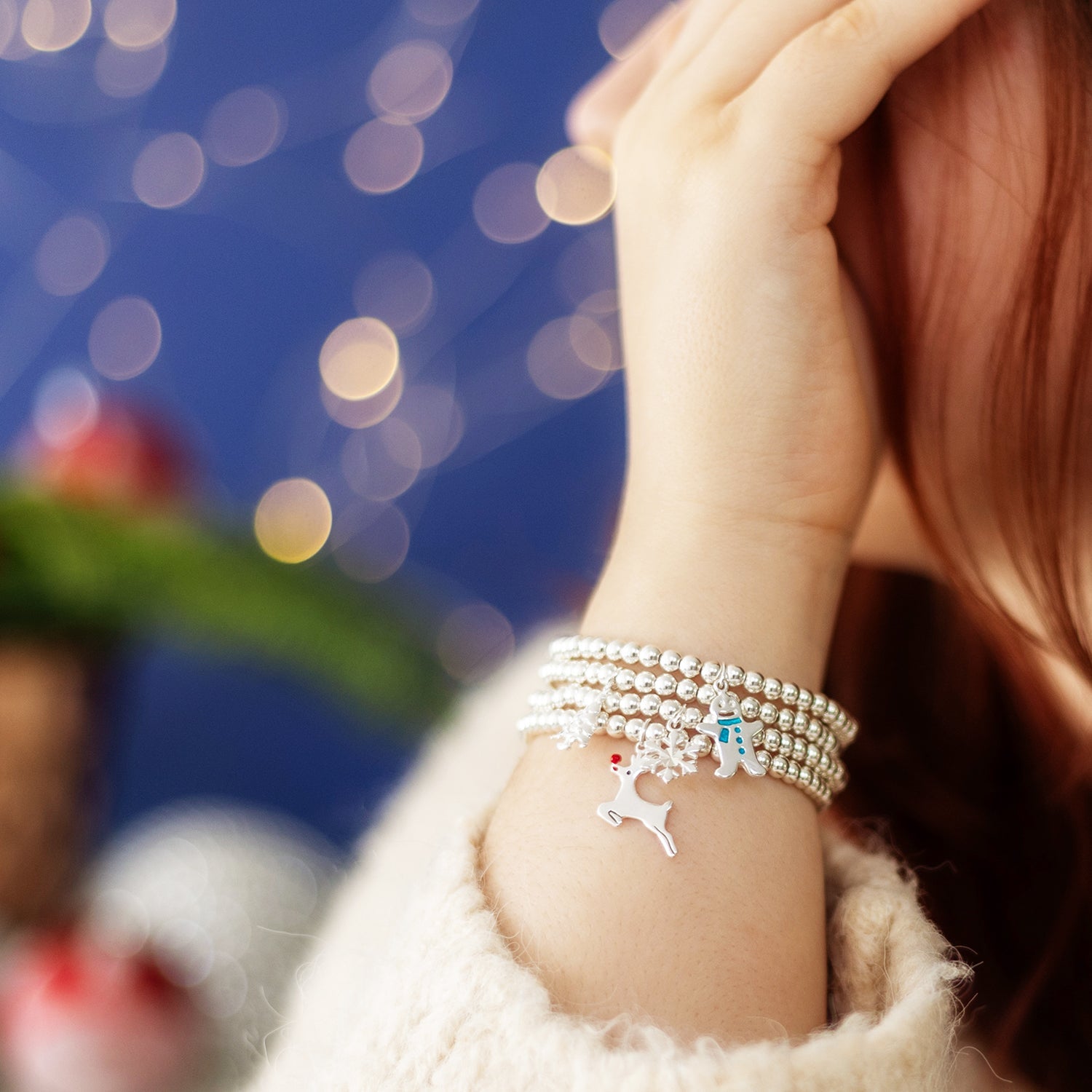  A person with long hair is wearing several silver beaded bracelets featuring charms, perfect as a Christmas jewellery gift. The background is blurred with lights, with a festive atmosphere. The person is partially covering their face with one hand