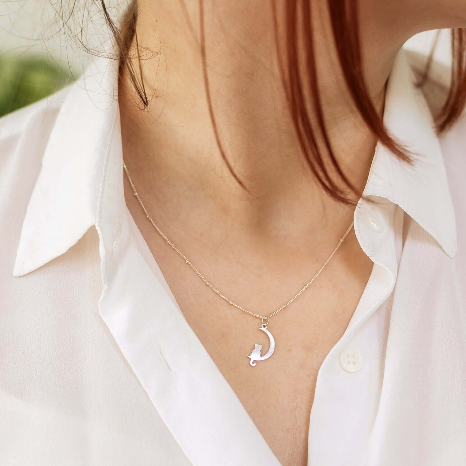 Close-up of a person wearing an open-collared white blouse, showcasing a delicate necklace. The necklace features a pendant in the shape of a crescent moon with a cat sitting on it. Hair partially obscures the viewer's left side of the necklace.