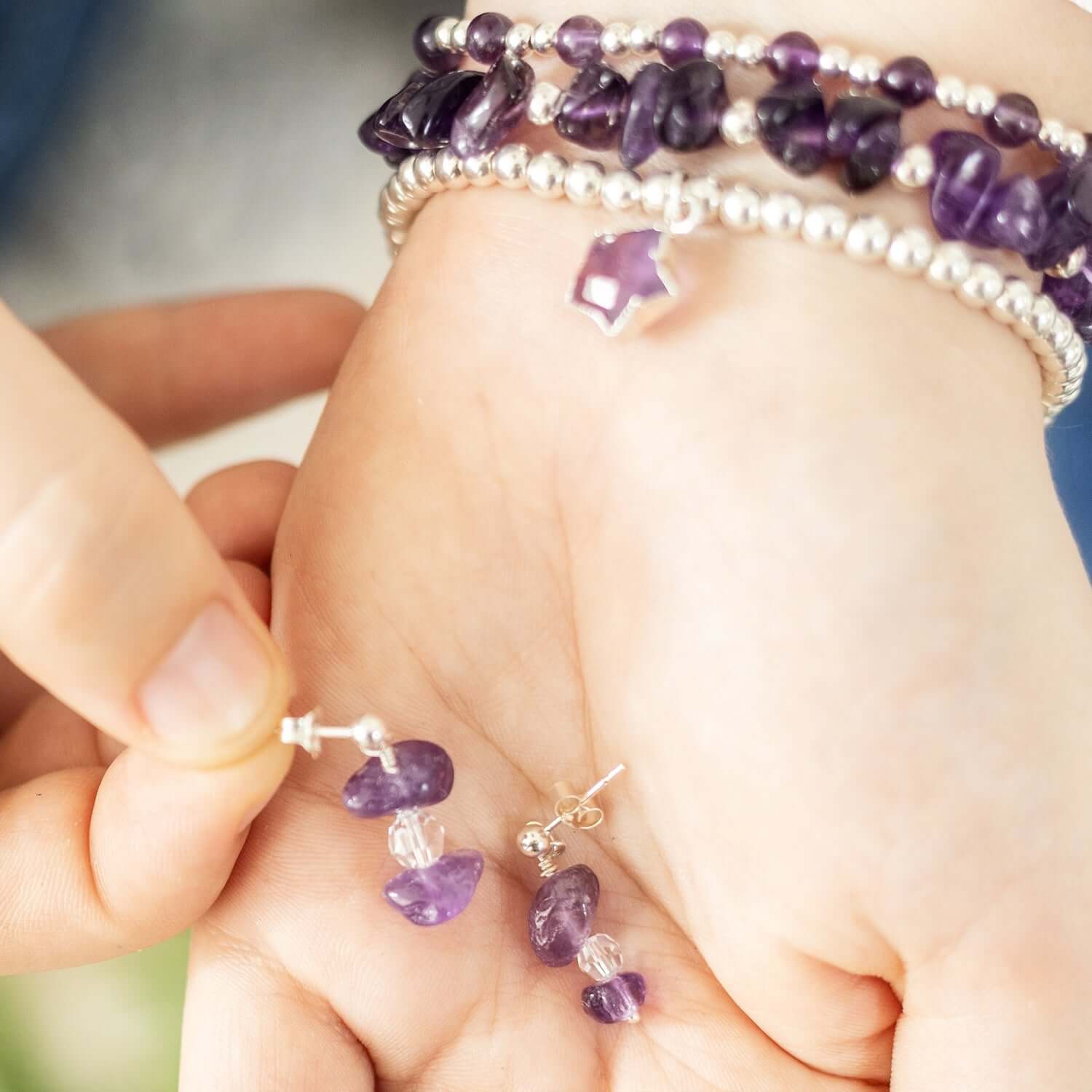 Close-up of a person holding a pair of gemstone drop earrings made with purple beads. The hand is wearing matching bracelets with similar purple stones and silver beads. The background is blurred, focusing attention on the jewelry.