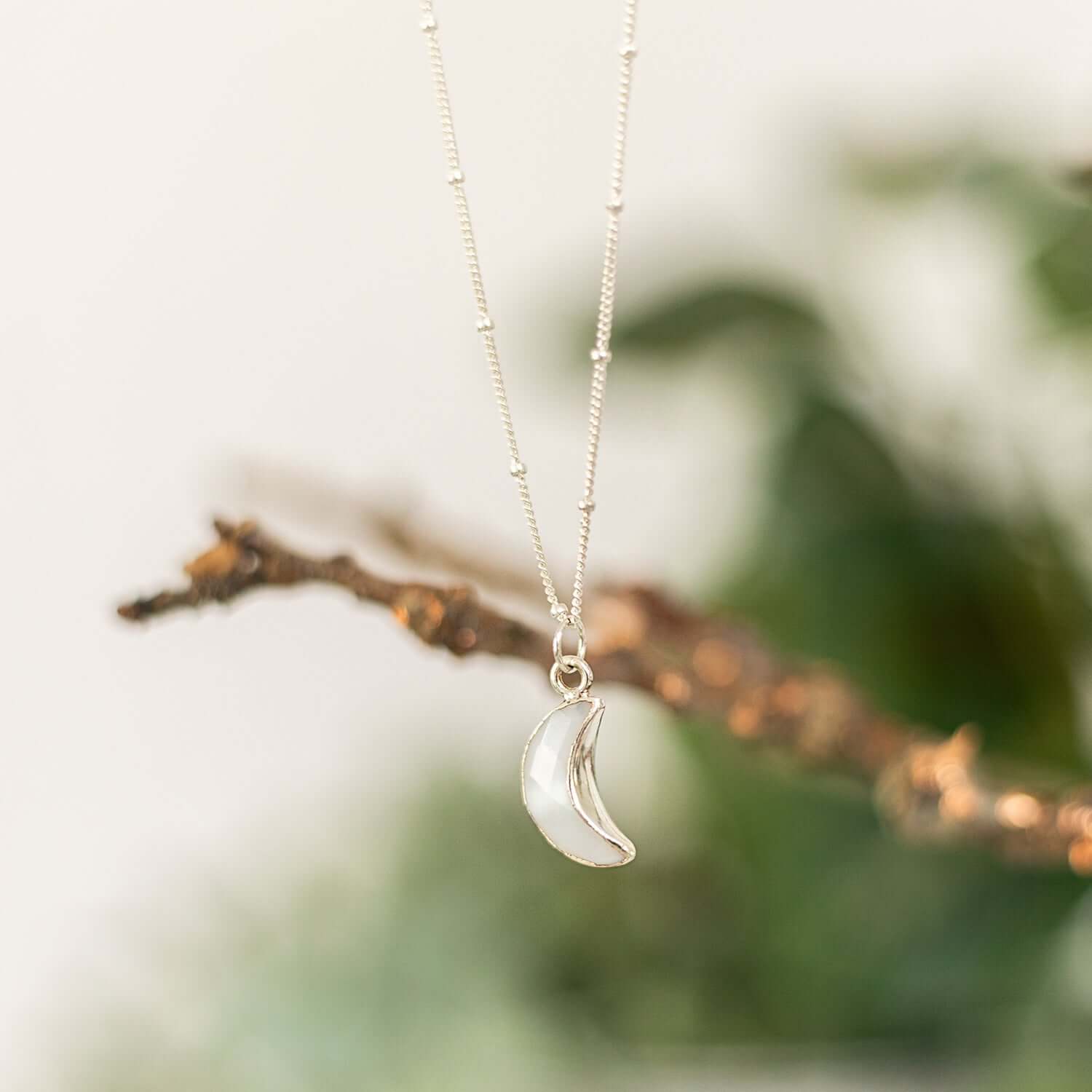  A delicate womens sterling silver necklace with a small crescent moon pendant hangs from a thin beaded chain. The background is softly blurred, featuring green foliage, creating an elegant and serene setting for the jewellery