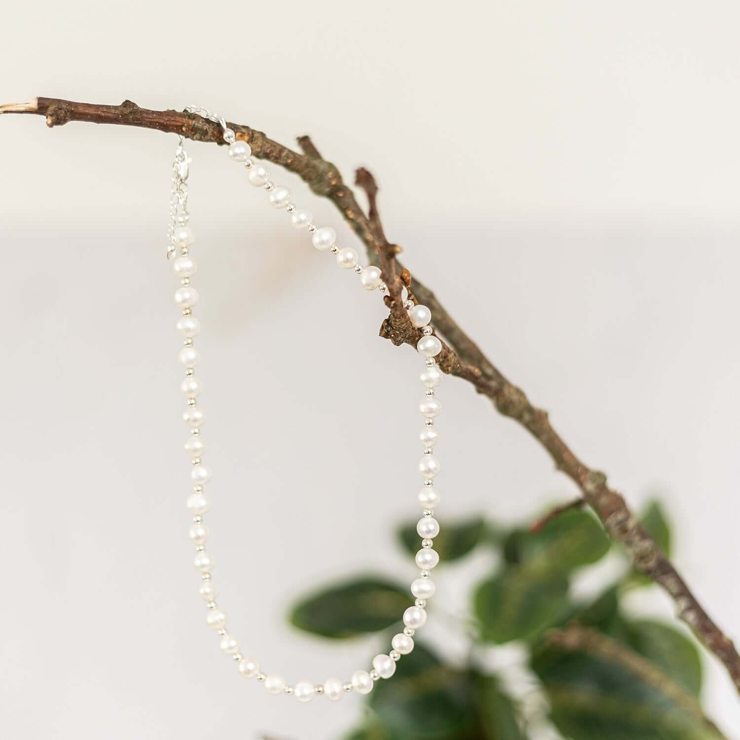  A pearl necklace with small, evenly spaced pearls is draped over a branch. The background is softly lit and slightly blurred, with hints of green leaves visible, giving the image a natural and aesthetic reminiscent of beaded necklaces UK collections.