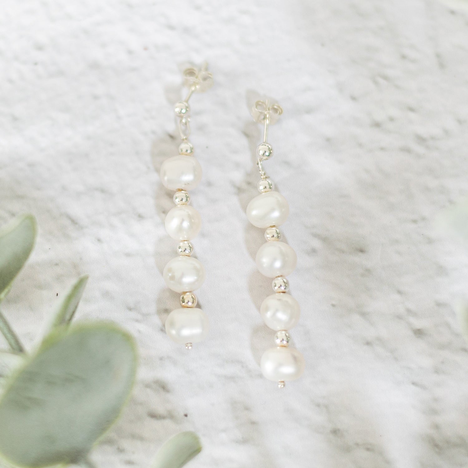  A pair of silver pearl drop earrings featuring a vertical arrangement of small white pearls interspersed with silver bead accents. The earrings are laid on a light surface, accompanied by green leaves on the left corner, creating a delicate aesthetic
