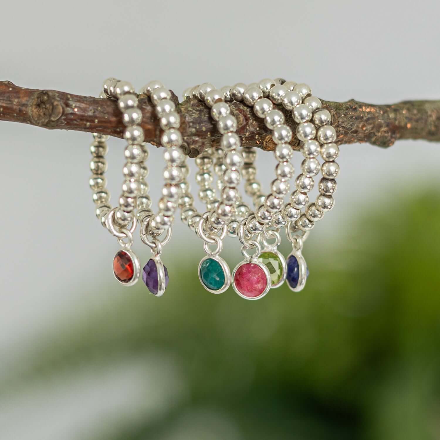 A group of silver beaded rings with colorful gemstone charms, representing birthstone jewellery uk, hanging on a tree branch. The rings feature gemstones in various colors, incl red, purple, green, pink, and blue, against a blurred green background