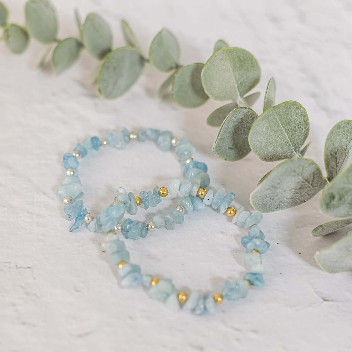  Two aquamarine gemstone bracelets, perfect as birthstone jewellery for March, are adorned with small gold and silver beads. They are displayed on a white surface with fresh green eucalyptus leaves arranged decoratively behind them