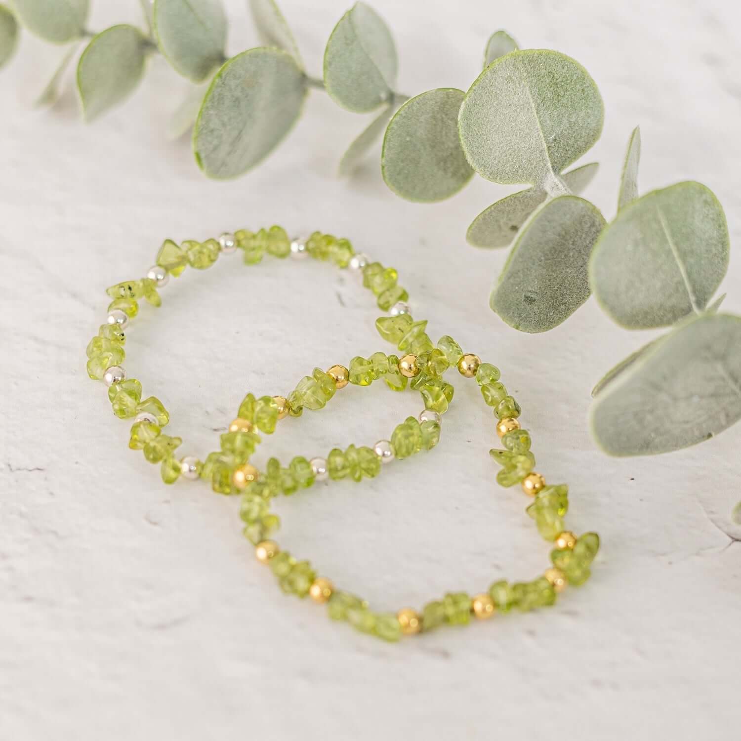 Two green peridot bracelets,featuring beads intermixed with small beads are placed on a light-colored surface.Highlighting August birthstone jewellery, a branch with round, silvery-green leaves serves as a backdrop, adding a natural element to the display