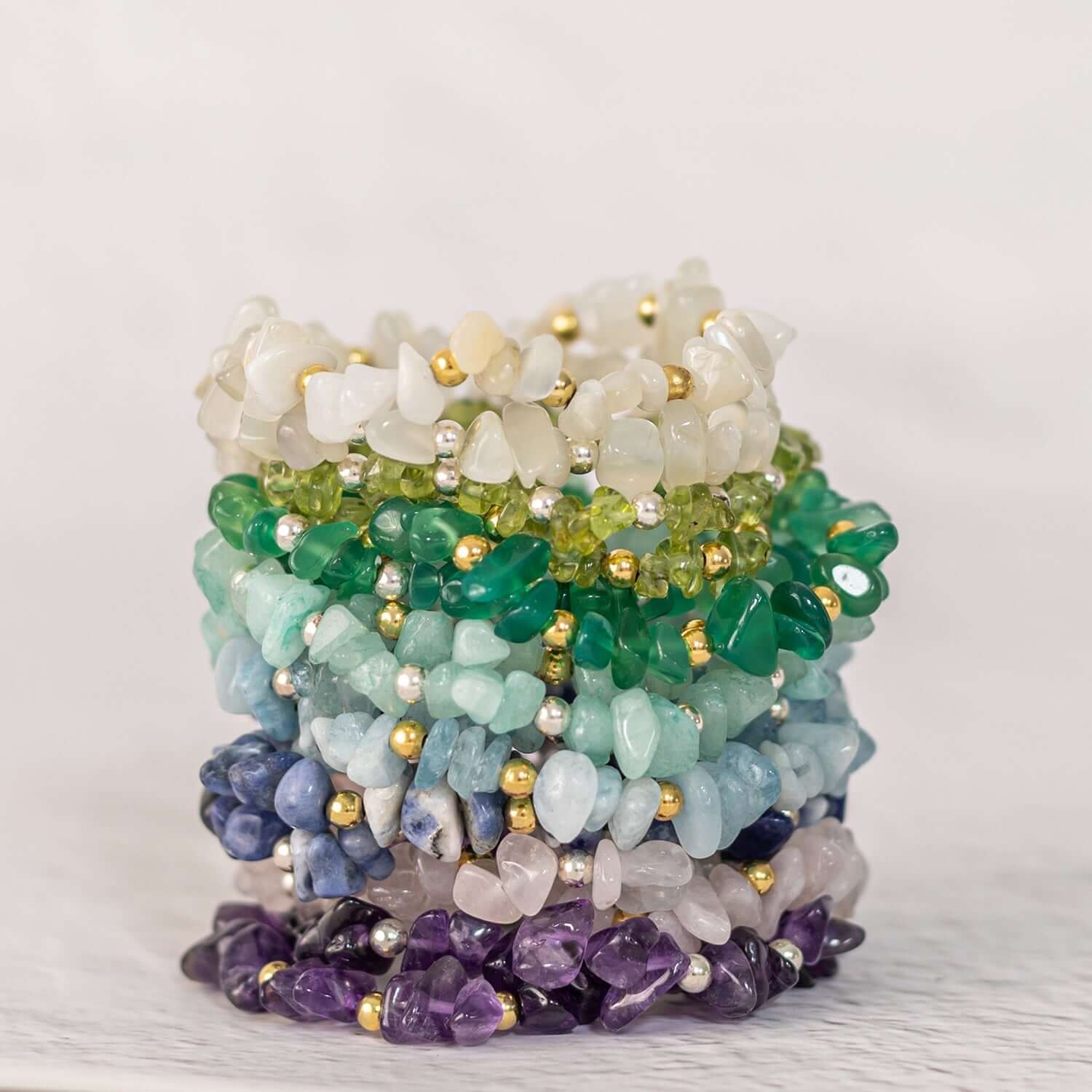 A stack of beaded gemstone bracelets featuring various colors: white, green, blue, and purple. Each bracelet is composed of irregularly shaped polished stones with small gold and silver beads, womens gold jewellery, arranged neatly on a light background
