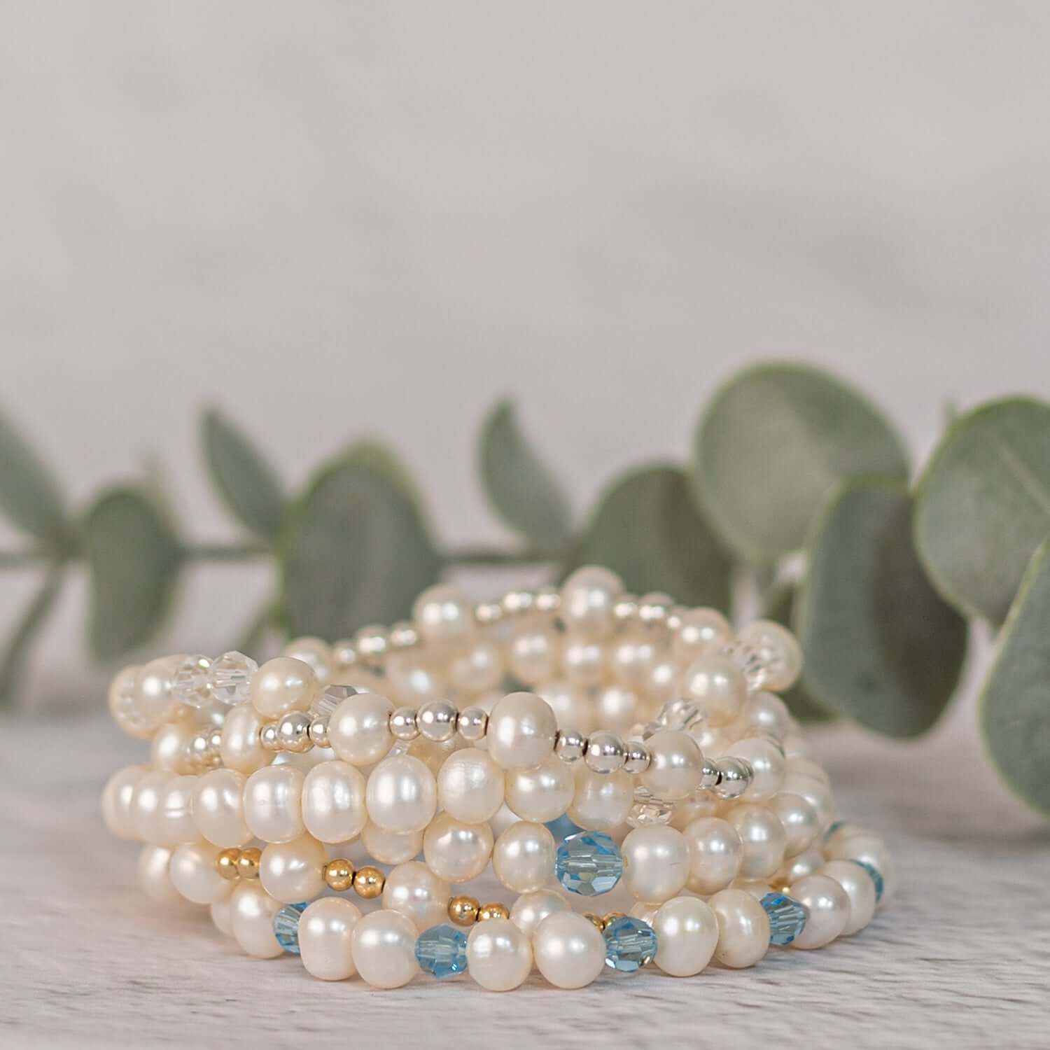 A multi-strand women's pearl bracelet made of white pearls, gold and silver beads, and blue crystals is coiled in a circular shape. The bracelet is set against a soft-focus background of green eucalyptus leaves on a light-colored surface