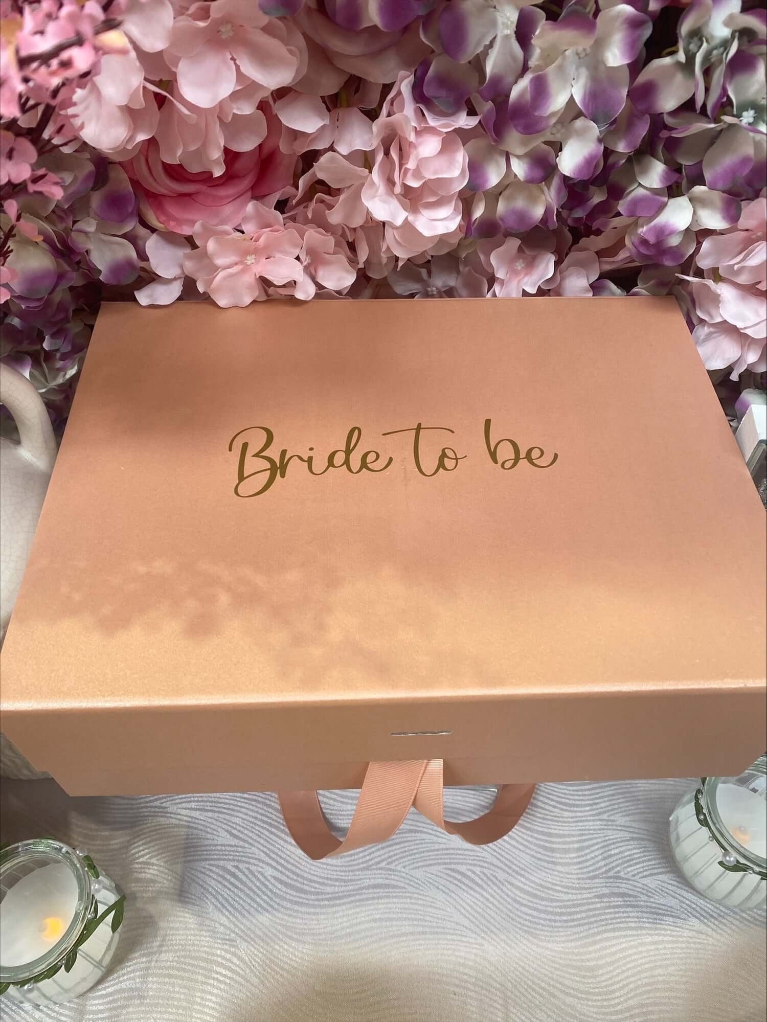  A pink gift box with "Bride to be" written in gold script on the lid is surrounded by a floral arrangement of pink and purple flowers. The personalised gift box is tied with a matching pink ribbon and displayed on a white surface