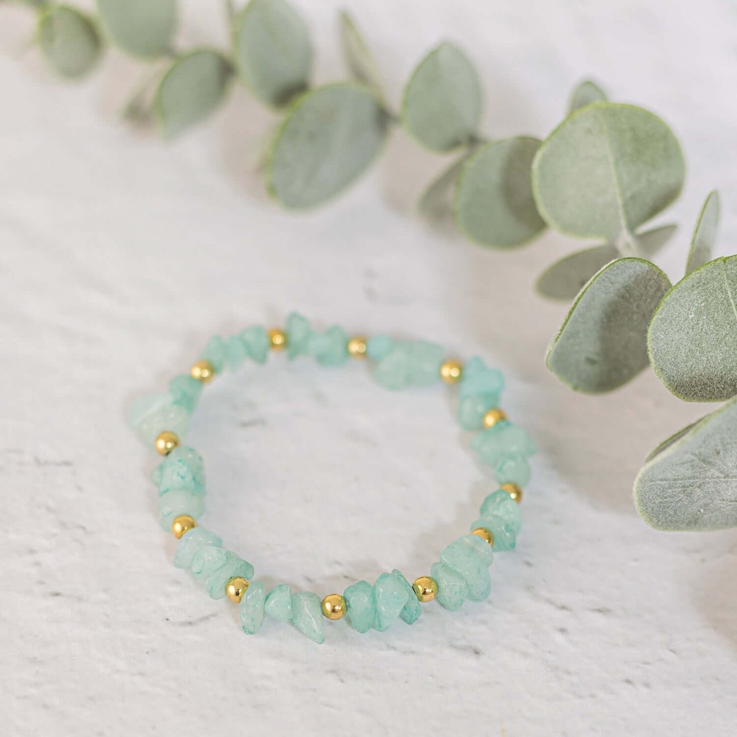 A delicate Made Here with Love Amazonite Gemstone Bracelet made of light green chips alternating with small gold beads is placed on a light gray surface. The bracelet, adorned with semi-precious gemstones, is accompanied by a sprig of eucalyptus leaves in the background, adding a natural touch to the scene.