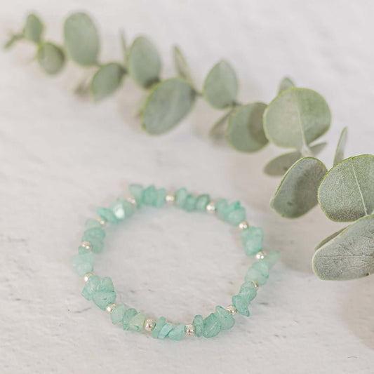 A delicate Amazonite Gemstone Bracelet by Made Here with Love made of small, irregular light green stones and tiny sterling silver beads is arranged on a white surface. Nearby, there is a branch adorned with several eucalyptus leaves, adding a touch of natural elegance to the composition.