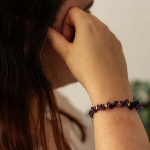 A person with long dark hair, resting their chin on their hand, showcases a handcrafted bracelet made of small purple and gold beads. The background is blurry, focusing attention on the Amethyst Crystal Bracelet by Made Here with Love and part of the person's face and hair.