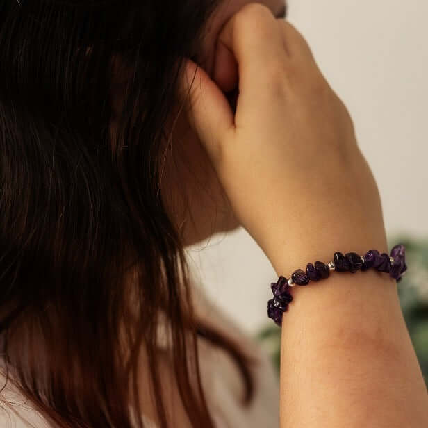 A person with long dark hair is seen from the side, with their hand placed near their face. They are wearing an Amethyst Crystal Bracelet by Made Here with Love, showcasing beautiful February Gemstone beads on the wrist of the raised hand.