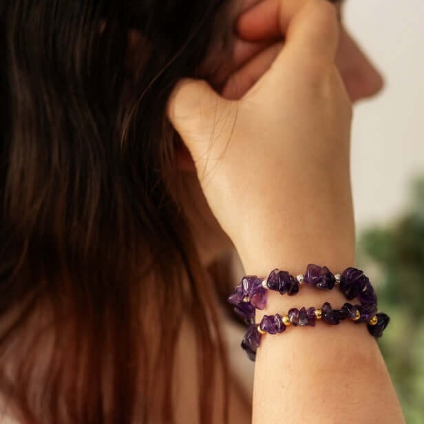 A person with long, dark hair is seen from the back with their hand touching their hair. They are wearing two handcrafted Amethyst Crystal Bracelets from Made Here with Love on their wrist, showcasing the beautiful February Gemstone, with blurred greenery in the background.