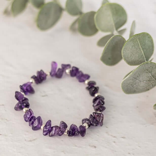 A handcrafted Amethyst Crystal Bracelet by Made Here with Love, adorned with irregularly shaped amethyst stones and small silver beads, is displayed on a white surface. Green eucalyptus leaves are positioned around the bracelet, providing a natural backdrop. Perfect for those who cherish February Gemstone elegance.