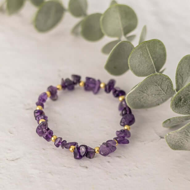 A delicate purple Amethyst Crystal Bracelet from Made Here with Love with small gold accents is displayed on a textured light surface, beside green eucalyptus leaves. This handcrafted jewellery piece beautifully showcases the February Gemstone.