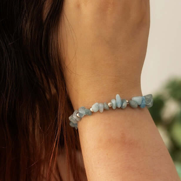 A close-up of a person's wrist adorned with an Aquamarine Stone Bracelet by Made Here with Love made of light blue irregularly-shaped handcrafted gemstones and small silver beads. The person’s long brown hair is visible and a blurred green plant is in the background, making it a perfect March Birthday gift.