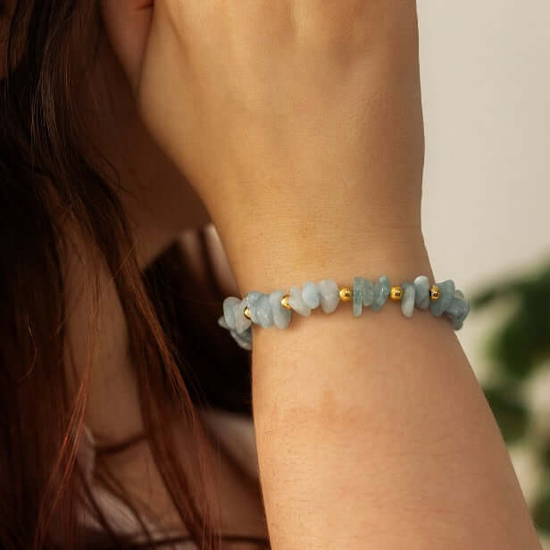 Close-up of a person wearing an Aquamarine Stone Bracelet from Made Here with Love, adorned with irregularly shaped light blue gemstone beads interspersed with small gold-colored beads. The handcrafted gemstones catch the light as the person's hand is raised near their face, with long brown hair partially visible. The background is neutral.