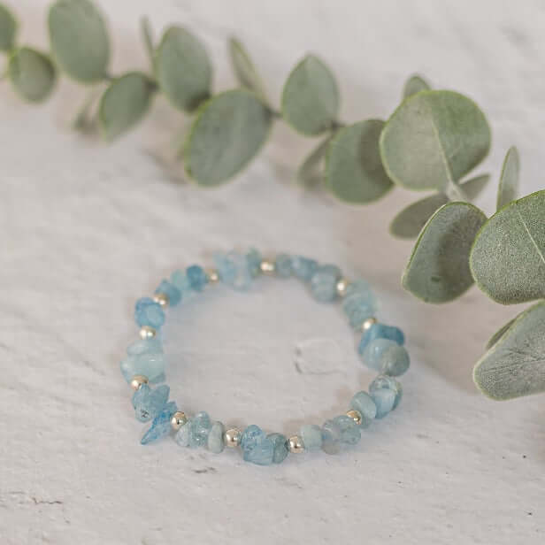 A Made Here with Love Aquamarine Stone Bracelet made of light blue gemstones and small silver beads rests on a light-colored surface. Next to it, there's a sprig of eucalyptus leaves. The soft lighting creates a serene and elegant atmosphere, making it the perfect March birthday gift.