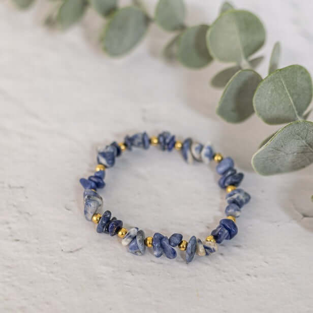 A Sodalite Crystal Bracelet by Made Here with Love, featuring irregular blue stones and small gold beads, is placed on a light-colored surface. Green eucalyptus leaves partially encircle the bracelet, adding a touch of nature to the scene.