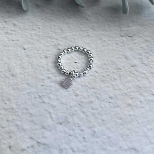 A Made Here with Love Rose Quartz Birthstone Ring features intricate silver beads and a small circular charm, elegantly placed on a light gray textured surface. Blurry green leaves in the upper part of the background highlight the sophistication of this Sterling Silver piece.