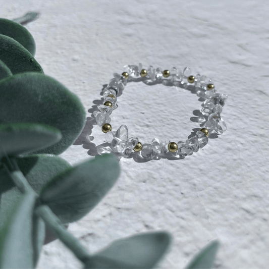 A stunning Clear Quartz Bracelet by Made Here with Love, featuring clear, irregular-shaped semi-precious gemstones and small, round, golden beads, lies elegantly on a textured white surface. Sterling silver beads add a touch of sophistication. Green leaves on the left side enhance this well-lit composition.