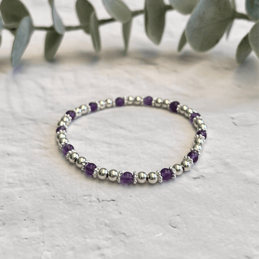 An Amethyst Stone Bracelet by Made Here with Love, adorned with a delicate mix of alternating shiny sterling silver beads and deep purple accents, is elegantly arranged in a circle on a light gray surface. The background includes softly focused green leaves, introducing a natural touch to the image.